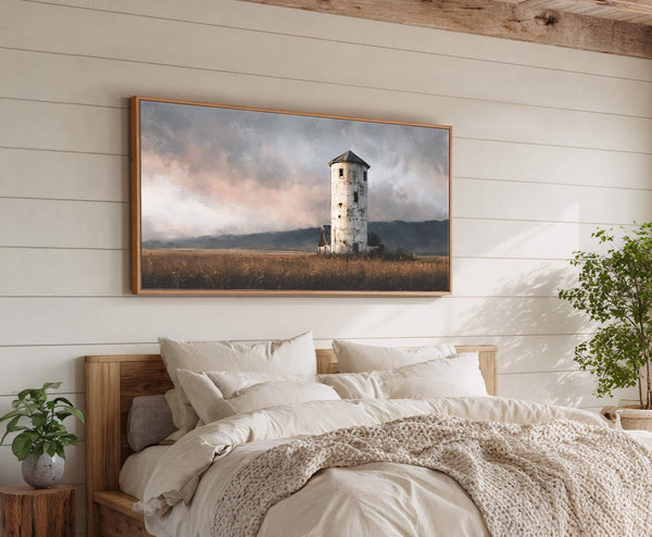 This horizontal canvas wall art features a rustic painting of a weathered tower in a field under a dramatic sky, framed in natural wood and hung above a bed on.
