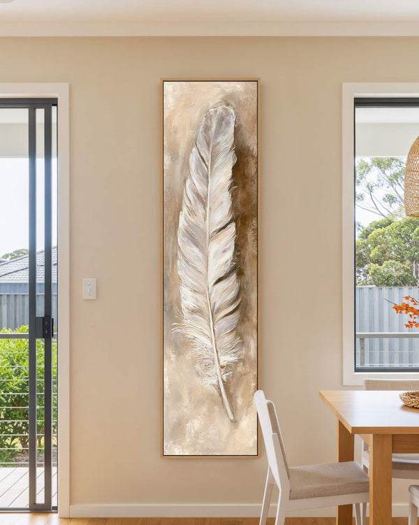 This vertical art print features a textured feather in a neutral palette, displayed in a light oak floater frame on a beige dining room wall near a wooden table.