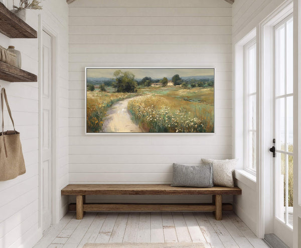 A horizontal, impressionistic landscape painting print of a country road winding through a field with scattered wildflowers, framed in white and displayed on a white paneled wall above a wooden.