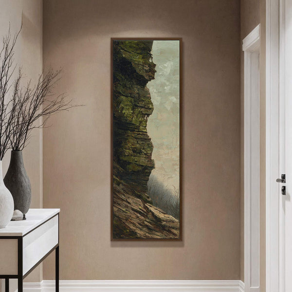 This vertical canvas wall art features a dramatic, painterly depiction of a mossy rock face with a misty sky, framed in dark wood, displayed on a tan wall above a.