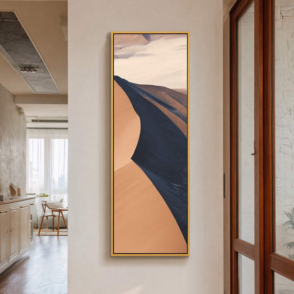 This vertical canvas wall art features abstract desert dunes in shades of tan, dark blue, and pale pink, framed in gold, displayed on a neutral wall beside a wooden door.