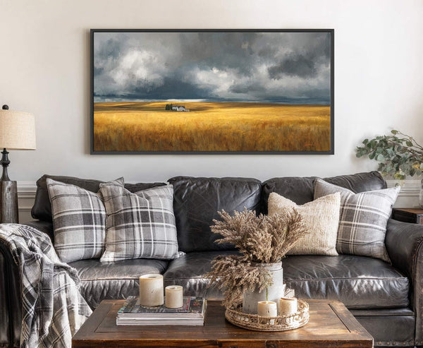 This horizontal canvas wall art displays a moody landscape with a golden field under stormy skies, framed in black, hanging above a dark leather sofa with plaid pillows in a.