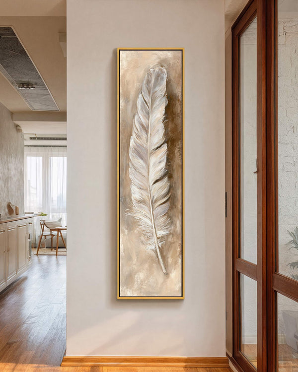 A vertical canvas print of a textured white feather in an earthy palette hangs in a gold floater frame on a white hallway wall beside a wooden window.