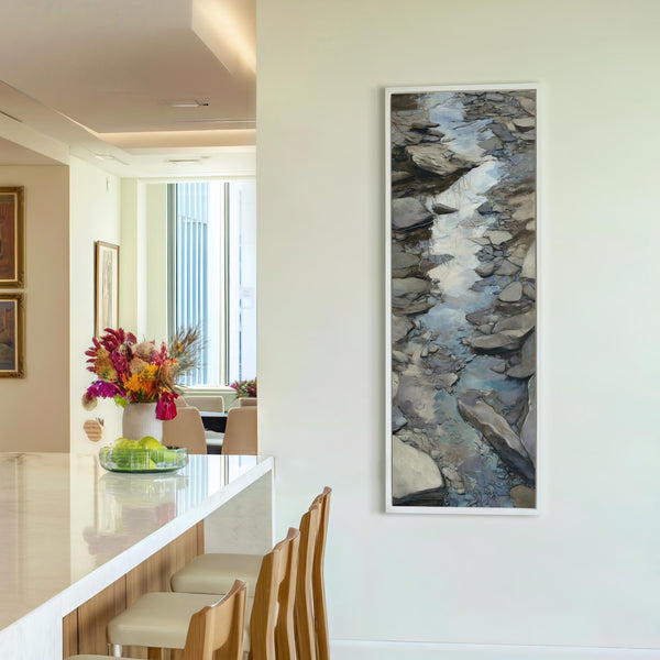 This vertical painterly art print features a rocky stream in earthy tones and is displayed in a white floater frame on a white wall beside a kitchen island.