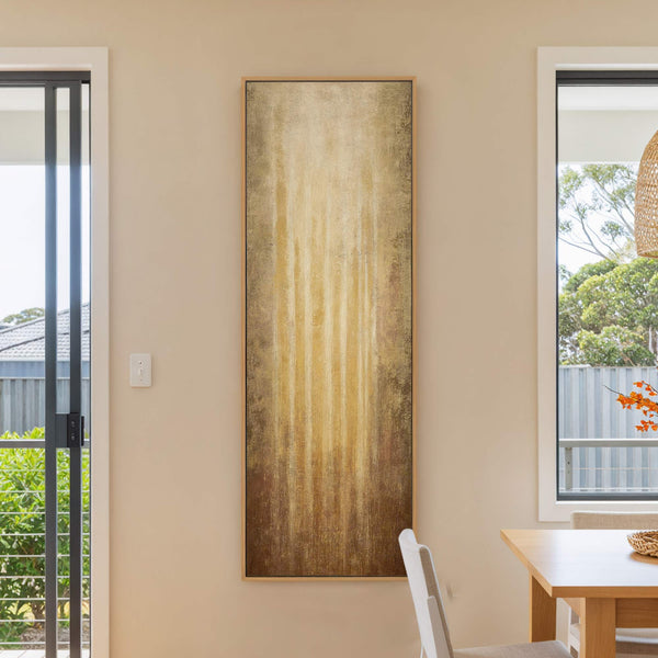 This abstract vertical canvas wall art print features a palette of rich gold and brown tones, with vertical brushstrokes creating a textured, atmospheric effect. It is displayed on a neutral.