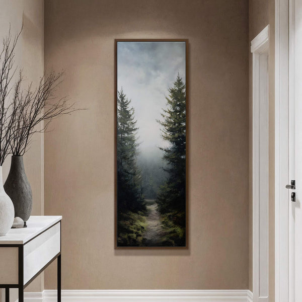 This vertical canvas wall art features a moody, atmospheric painting of a path through dark, misty pine trees. The art print is framed in natural wood and hangs on a.
