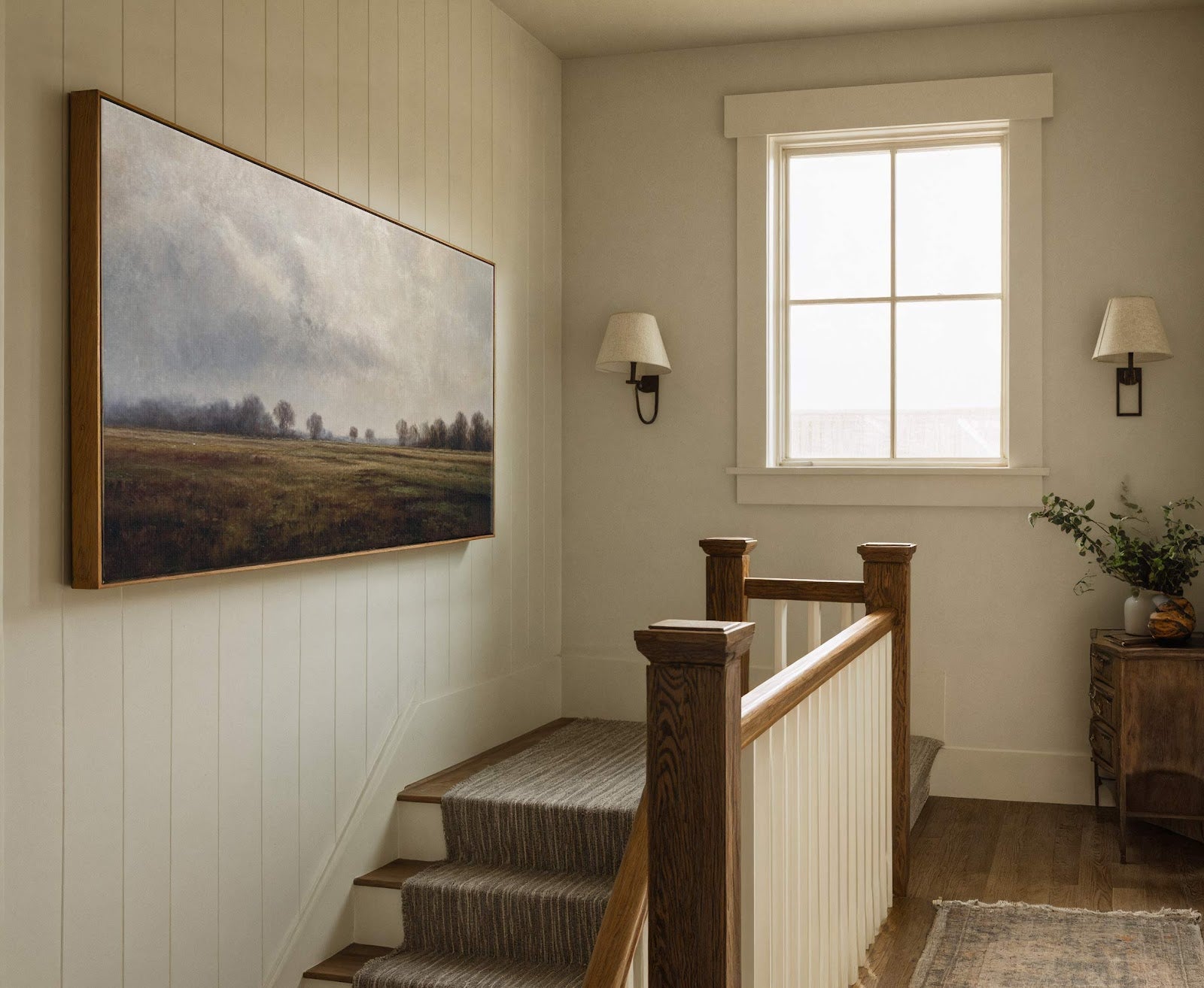 A horizontal canvas print features a subdued landscape with a muted palette of greens and grays, framed in natural wood, hanging on a paneled wall above a staircase with a.