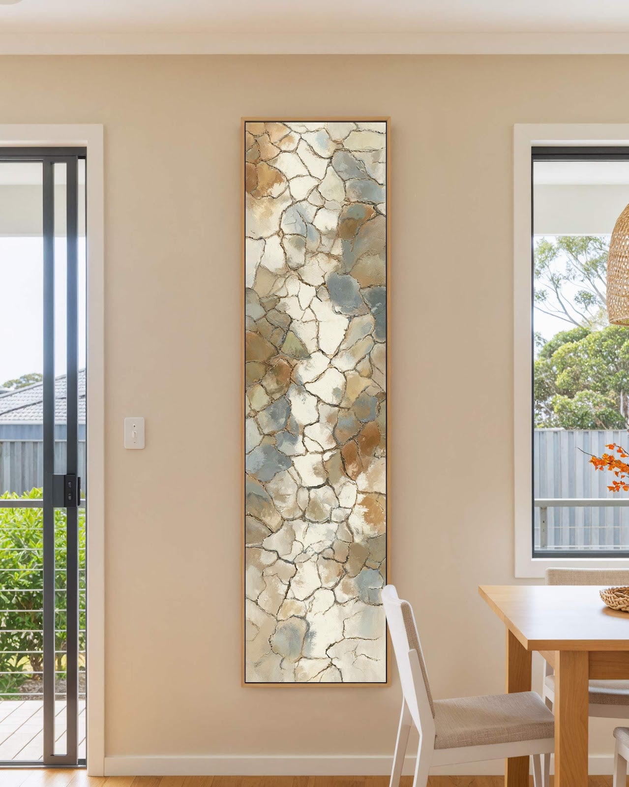 This vertical canvas print showcases an abstract earth-toned stone mosaic in a natural oak floater frame, hanging on a beige wall in a dining room near a wooden table.