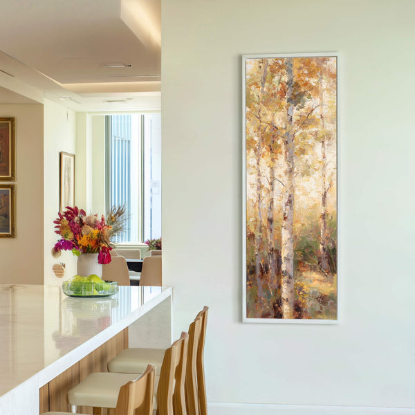 This vertical canvas wall art features an impressionistic forest scene with a warm, golden palette and is framed in white, displayed on a neutral wall with wooden bar stools visible.