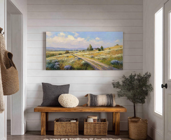 A horizontal canvas print captures a pastoral landscape with rolling hills, a dirt road, and scattered wildflowers under a blue, cloudy sky, displayed above a wooden bench in a room.