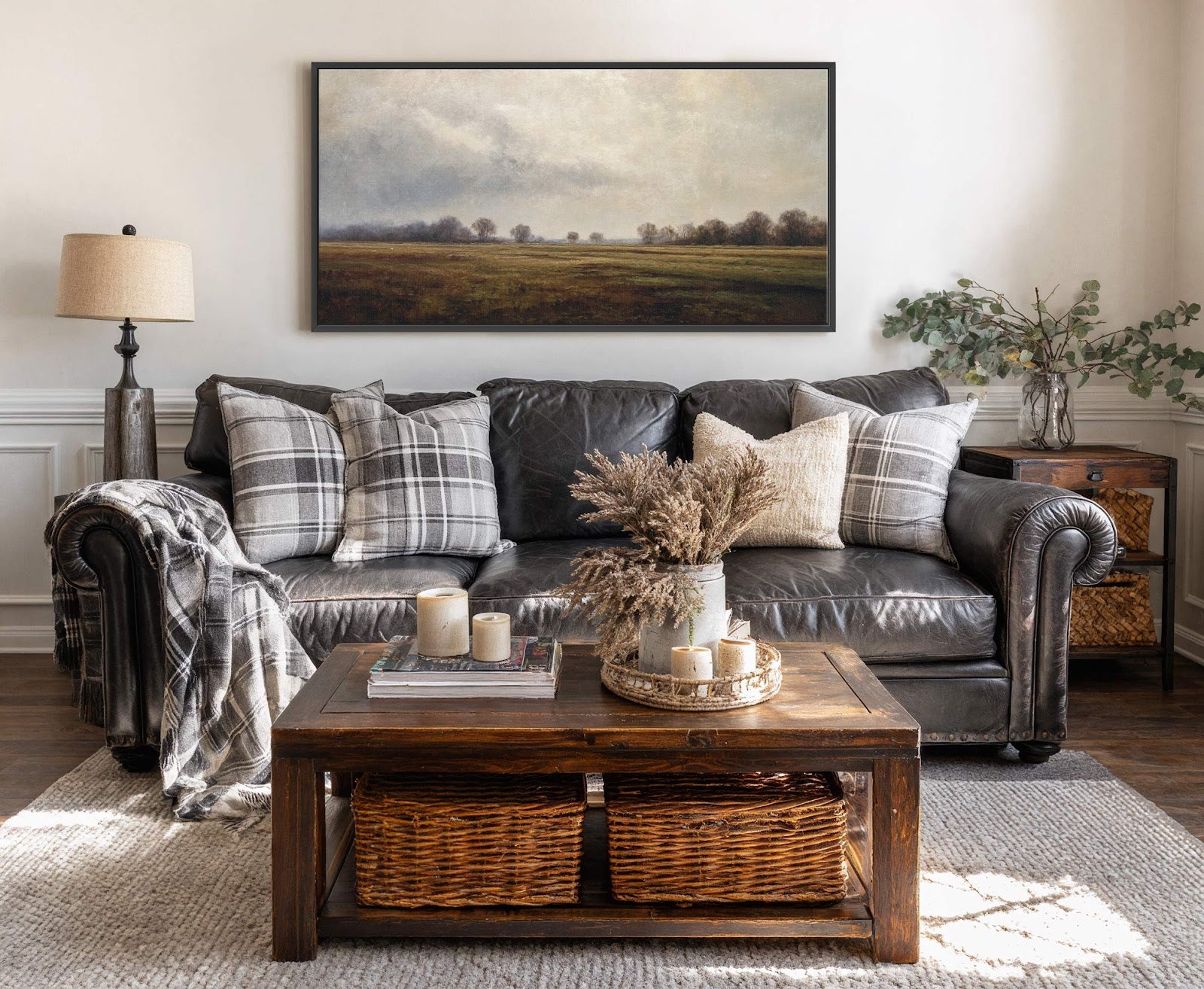 This horizontal canvas wall art features a misty landscape with muted greens and browns under a cloudy sky, displayed above a black leather sofa with plaid pillows and a dark.