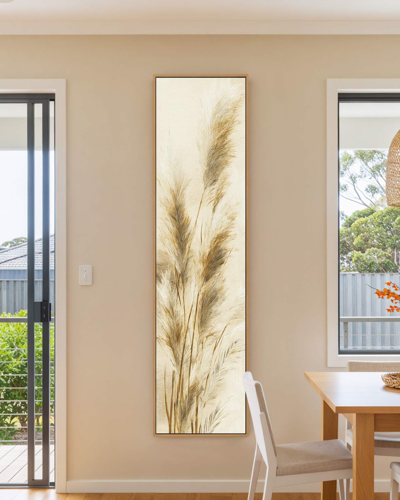 This vertical botanical art print showcases neutral pampas grass plumes in an oak floater frame, hanging on a beige wall in a dining room beside a wooden table.