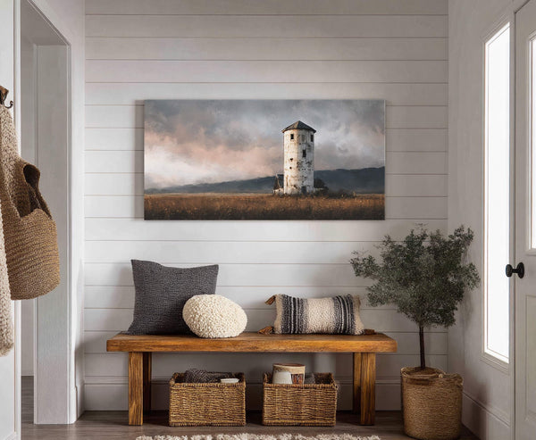 This horizontal canvas wall art depicts a weathered tower in a field under a cloudy sky, displayed above a wooden bench with pillows and baskets in a room with white.