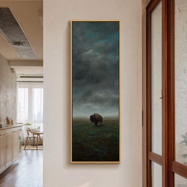 This vertical canvas print displays a lone bison in a moody, atmospheric landscape with a dark, stormy sky and a muted green palette, framed in gold and hanging on a.
