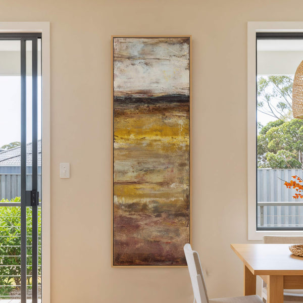 This vertical canvas wall art features an abstract landscape in earthy tones of yellow, brown, and white, with a textured appearance. It hangs on a beige wall between two windows.