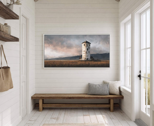 This horizontal art print displays a rustic, weathered tower against a dramatic, cloudy sky and a field of tall grass, framed in white and hung on a white paneled wall.