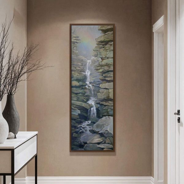 This vertical, framed canvas print depicts a naturalistic waterfall cascading between grey rock formations with muted colors and visible brushstrokes, displayed on a tan wall above a console table with.
