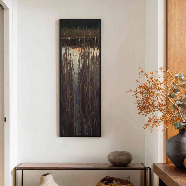 This vertical canvas wall art displays a moody impressionistic painting of reeds reflected in water with a dark palette and black floater frame, displayed in a room with white walls.