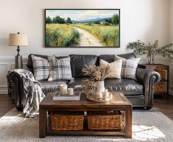 A horizontal canvas print displays an impressionistic landscape painting with a dirt road winding through a sunlit meadow of wildflowers, framed in black. The art hangs above a dark leather.