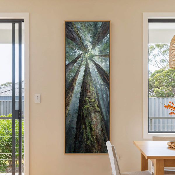 A vertical canvas print in a natural wood frame depicts a dramatic, upward-looking view of redwood trees with lush green moss, presented on a neutral wall next to a window.