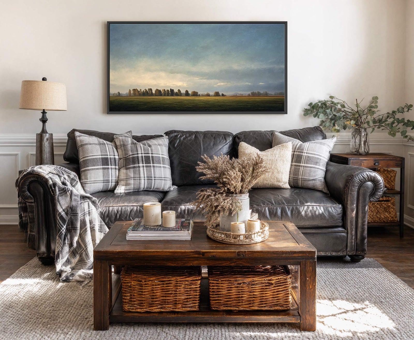 This textured landscape art print with a muted blue and green palette is a horizontal canvas in a black floater frame, displayed above a dark leather sofa on a light.