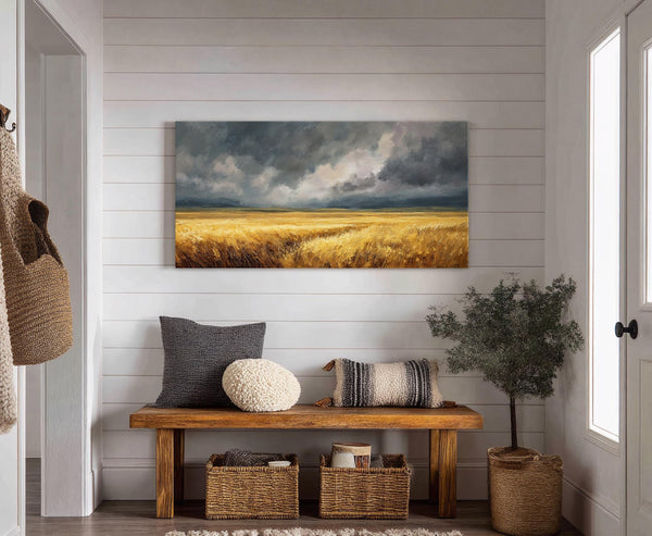 This horizontal, stretched canvas wall art depicts a golden wheat field under a dramatic, cloudy sky in a loose, painterly style, hung above a wooden bench with decorative pillows and.
