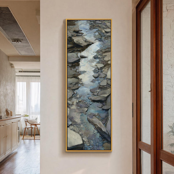 This vertical canvas wall art features a painterly rocky stream in earthy tones and blue, displayed in a gold floater frame on a white hallway wall near a wooden cabinet.