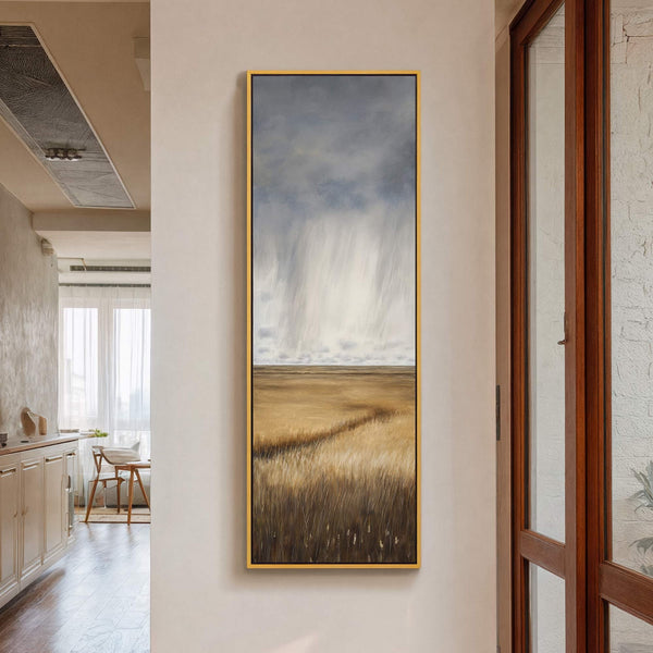 This vertical canvas print depicts a stormy sky over a golden field in a realistic style. It is displayed on a neutral wall next to a wooden door frame, with.