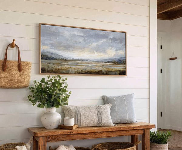 This horizontal canvas wall art features an abstract landscape in a muted palette of grays, blues, and earthy tones, framed in natural wood, displayed on a white paneled wall above.