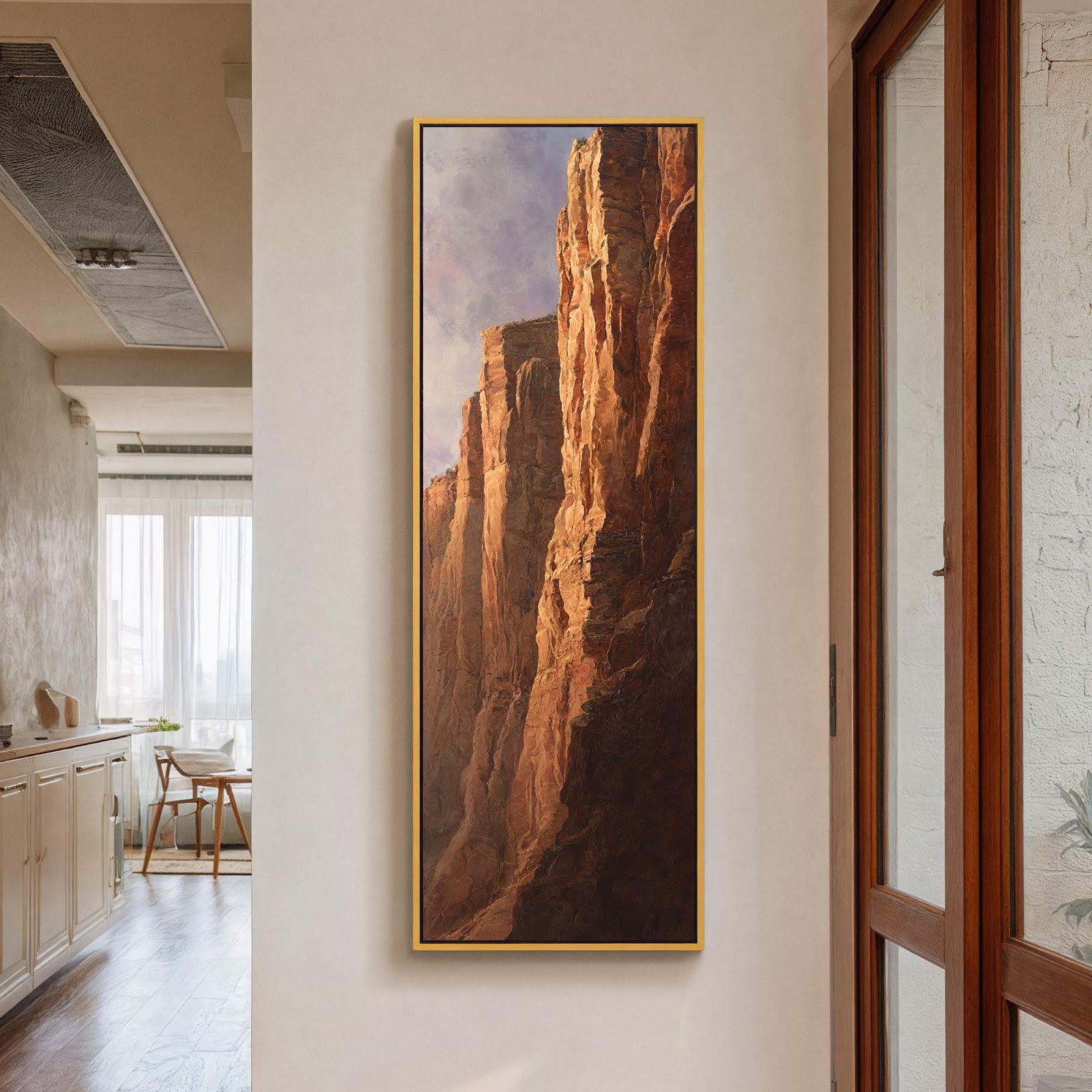 A vertical canvas wall art print depicts a dramatic desert canyon with warm, earthy tones and realistic style, framed in gold and hung on a light-colored wall in a room.
