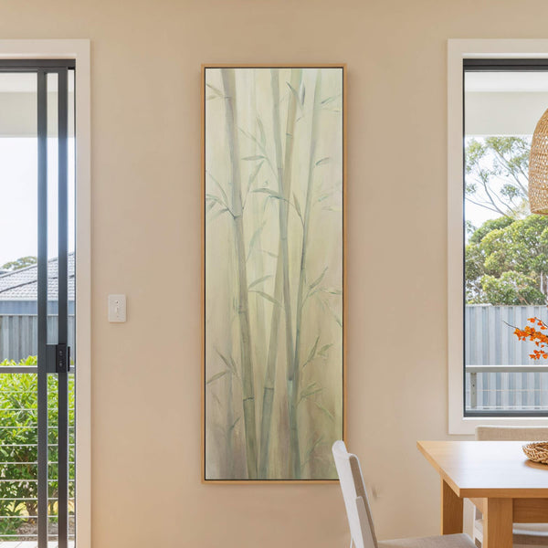 A vertical canvas print with a soft palette depicts bamboo stalks in a painterly style, displayed on a tan wall beside a sliding glass door.