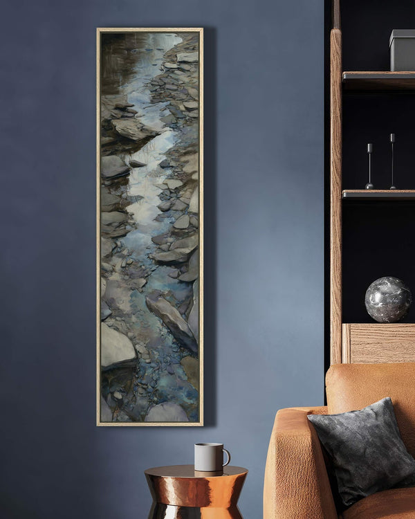 This vertical canvas print features an impressionistic rocky stream in cool tones, showcased in a natural wood floater frame on a dark blue wall beside a tan leather sofa.