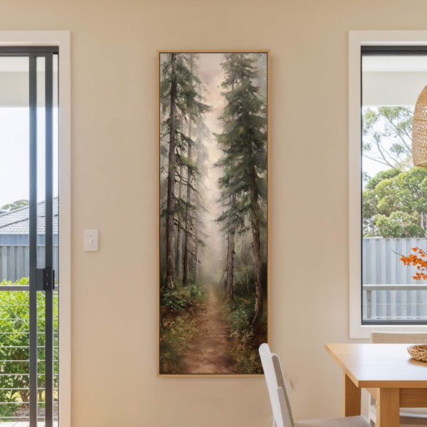 This vertical canvas wall art depicts a misty forest path with rich green and brown tones in a realistic style, hanging on a light beige wall near a window and.