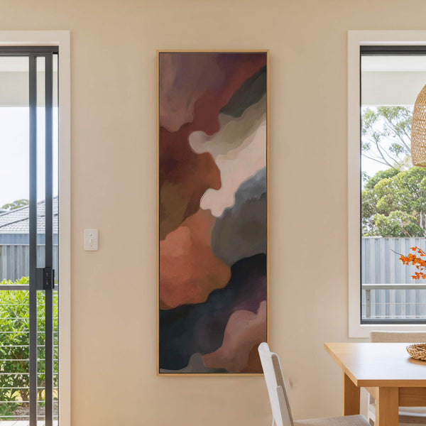This vertical canvas wall art features abstract shapes in a warm, earthy palette of rust, brown, and dark purple. It hangs on a light beige wall, with a window and.