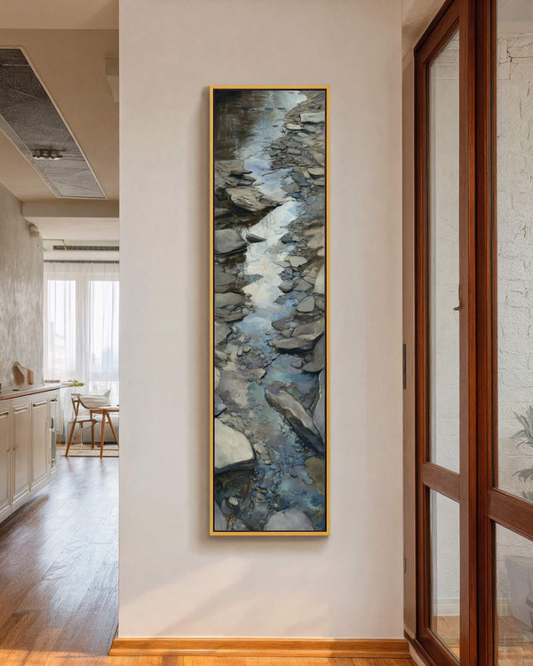 A vertical impressionistic canvas wall art of a rocky stream in cool blues and grays hangs in a gold floater frame on a white hallway wall near cabinets.