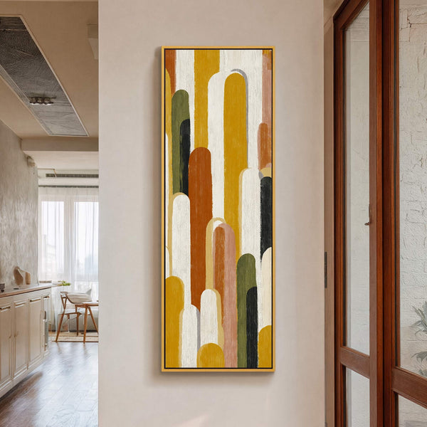 This vertical canvas print features an abstract geometric design with rounded shapes in a warm, earthy palette of yellow, orange, white, black, and green, framed in gold, hanging on a.