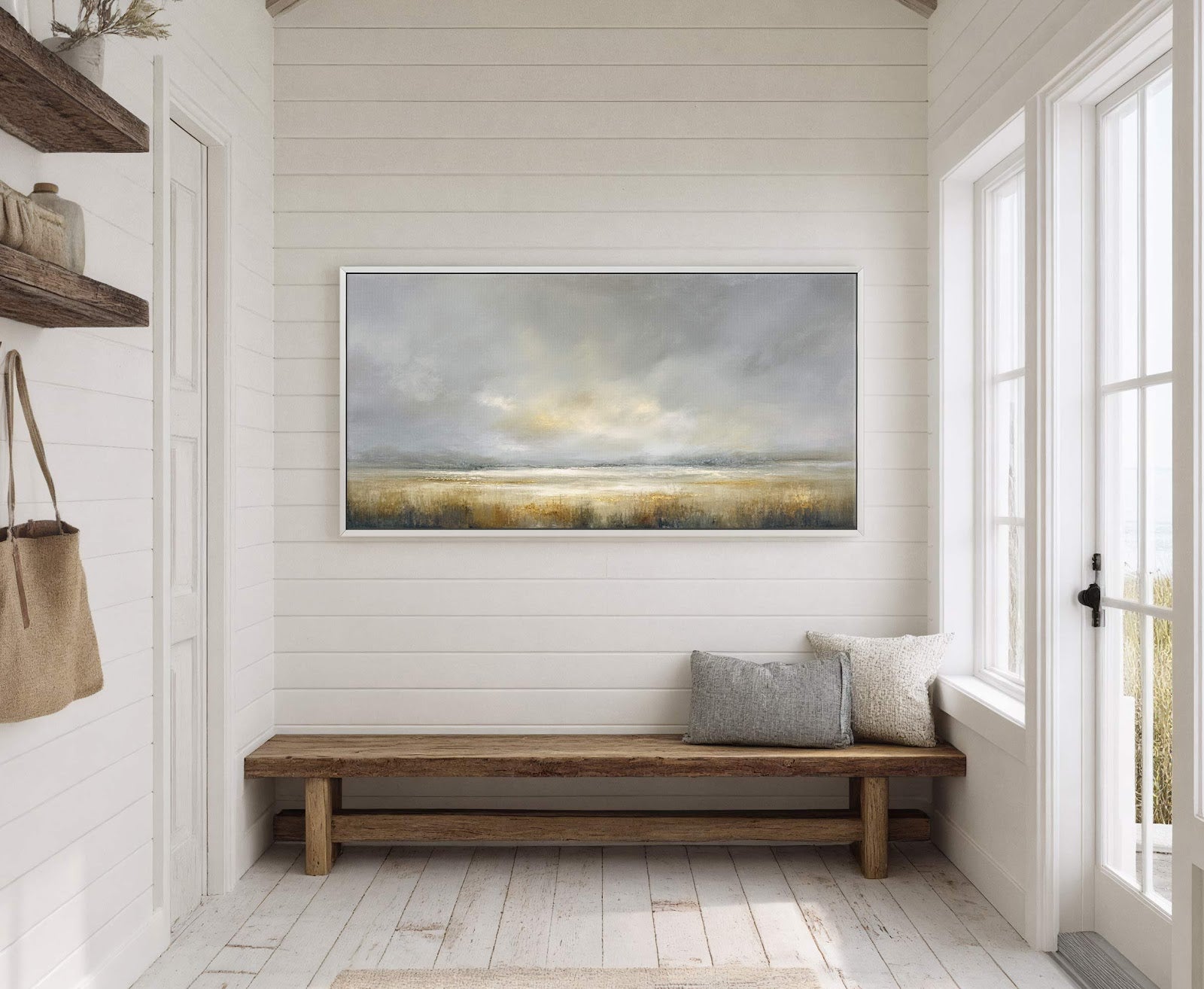 This horizontal canvas wall art features a muted palette abstract landscape with grassy fields under a cloudy sky, framed in white, hanging above a wooden bench in a room with.