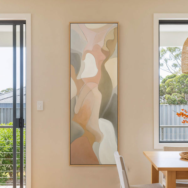 This vertical canvas wall art features abstract organic shapes in a soft palette of pink, beige, and gray, displayed on a neutral wall next to a window and a wooden.