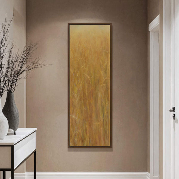 A vertical canvas print of a golden wheat field in a painterly style with warm earthy tones, framed in dark wood, hangs on a taupe wall above a white console.