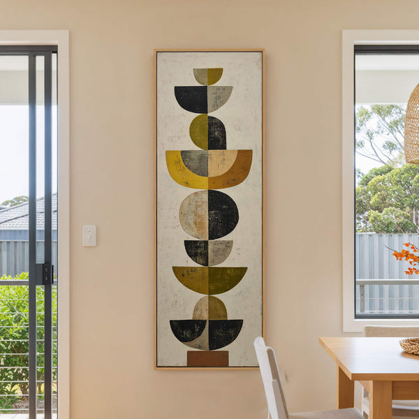 This vertical canvas print features an abstract geometric design with a palette of black, olive green, ochre, and brown, displayed in a natural wood floater frame against a cream wall.