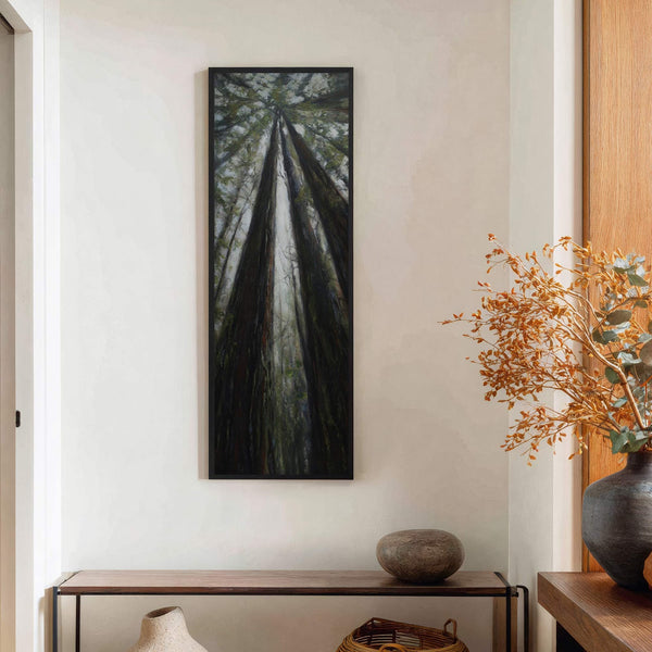 This vertical canvas wall art depicts tall trees looking upwards with a moody palette of dark greens and grays, set against a textured white wall above a console table.