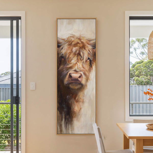 A vertical, framed canvas print of a Highland cow, rendered in a painterly style with a warm, earthy palette, is displayed on a neutral wall next to a sliding glass.