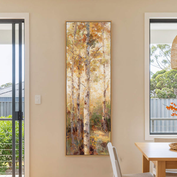 This vertical canvas print features an impressionistic forest scene with birch trees rendered in a warm palette of yellows, oranges, and greens, hanging on a beige wall next to a.