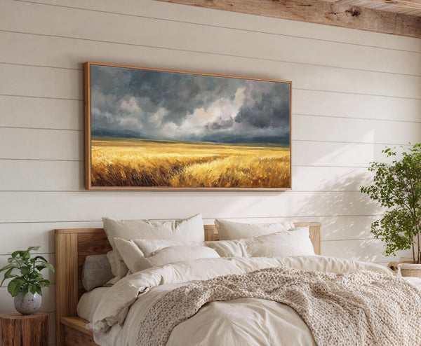 A horizontal canvas wall art print captures a golden field under a dramatic, cloudy sky in an impressionistic style, displayed above a bed with white linens in a room with.