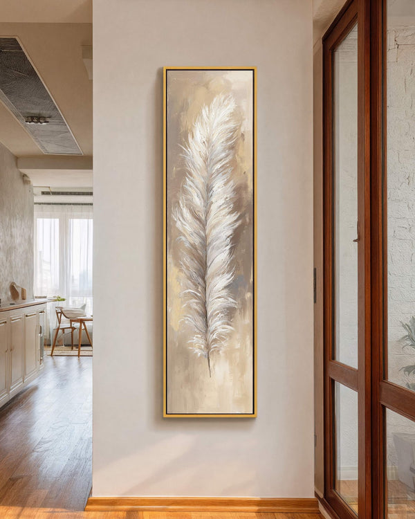 This vertical canvas wall art features an impressionistic white feather on a beige palette, displayed in a gold floater frame on a white hallway wall beside a window.
