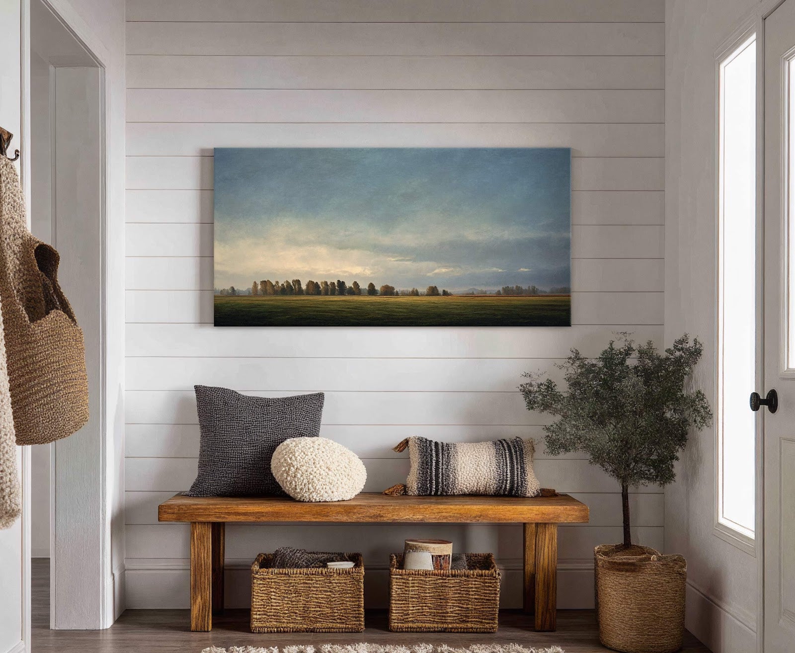 This horizontal canvas wall art displays a serene landscape with a distant treeline under a cloudy sky and soft, natural light. It hangs above a wooden bench with pillows and.