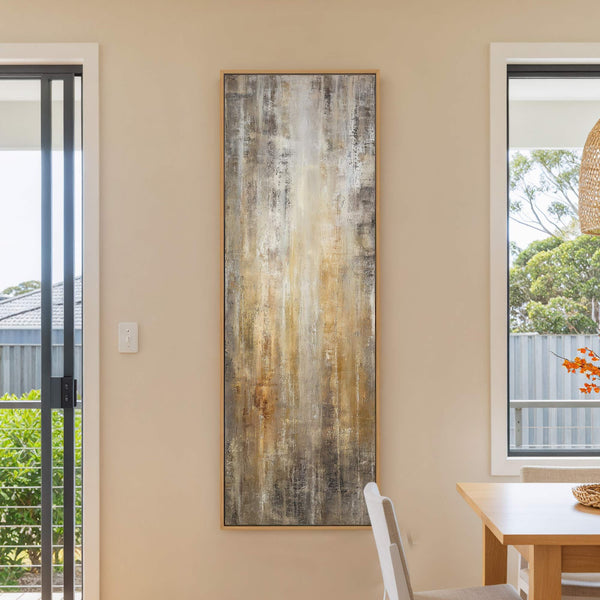 This vertical, abstract art print features a palette of brown, gold, and gray textures, framed in natural wood, and hangs on a beige wall next to a window and a.