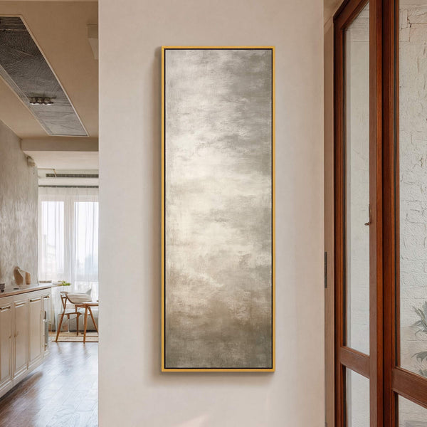 This vertical canvas wall art features an abstract impressionist style with a neutral palette of grays and whites, framed in a thin gold floater frame, displayed in a hallway with.