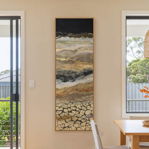 This vertical canvas wall art features an abstract landscape in earthy tones and gold, with a black textured top, framed in natural wood, displayed on a beige wall next to.