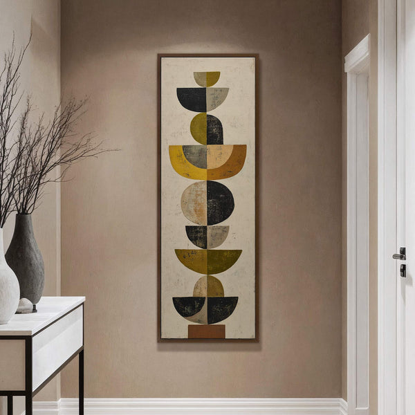 This vertical canvas wall art features an abstract geometric composition with a palette of mustard yellow, olive green, black, and gray, framed in walnut on a beige wall next to.